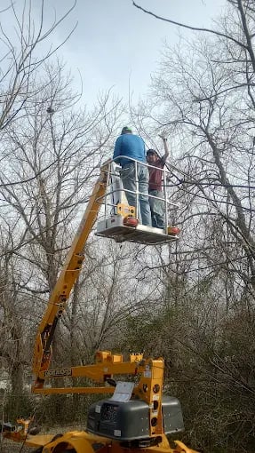 Worker in blue hard hat on yellow hydraulic lift among bare winter trees